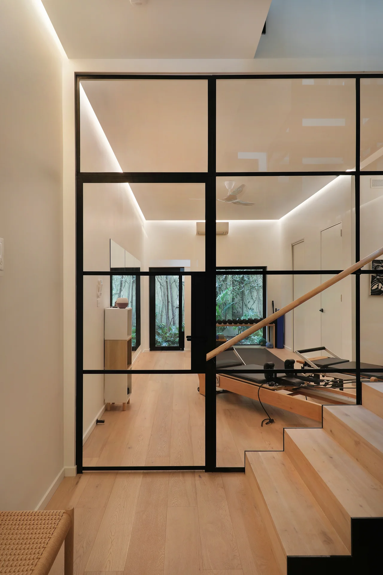 Black steel-framed glass partition wall looking into a modern Pilates studio, featuring light timber flooring, a custom timber staircase, and recessed downlights, George Street Fitzroy — Eco Building Design